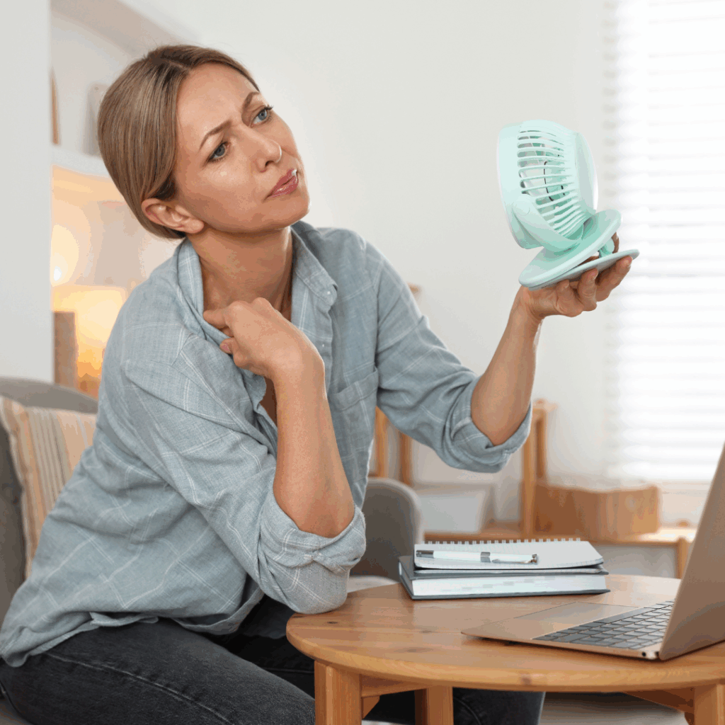 A midlife woman cooling herself with a small fan while working, showing discomfort from hormonal symptoms.